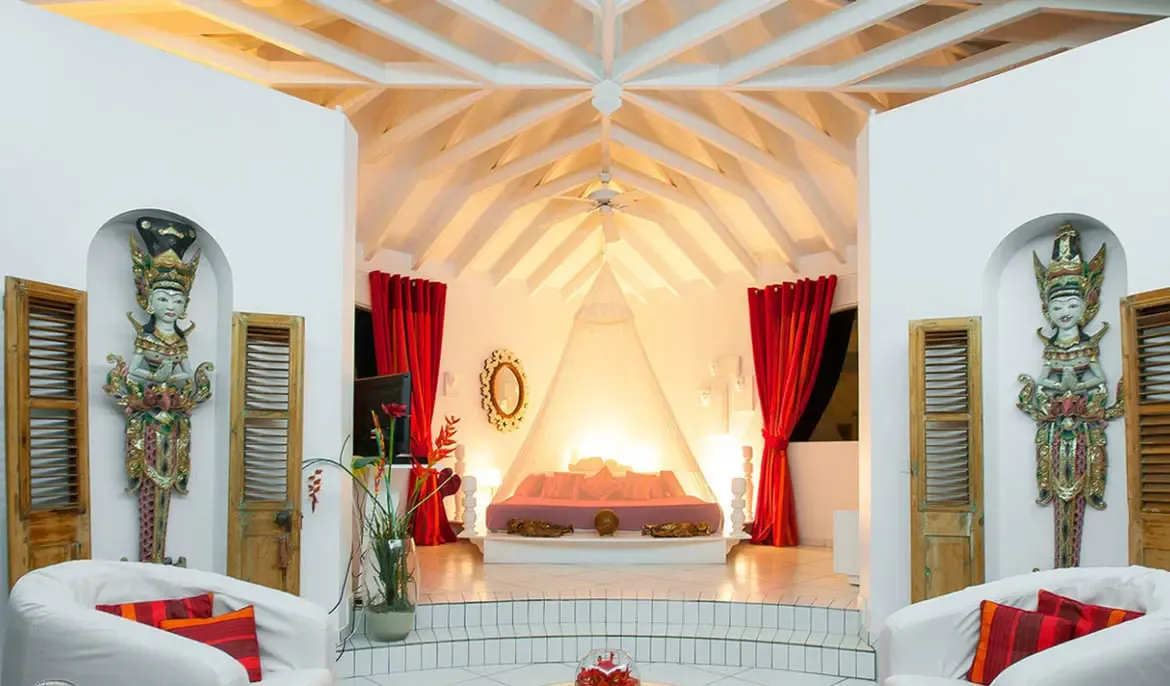 Retreat to the intimate sanctuary of Villa Byzance's bedroom, where a softly lit canopy bed anchors a space blending tropical elegance with cultural artistry. Rich red drapery, handcrafted Balinese statues, and vaulted ceilings create a luxurious escape in the heart of St. Barths.                                                                                                                                                                                                                                 +
 