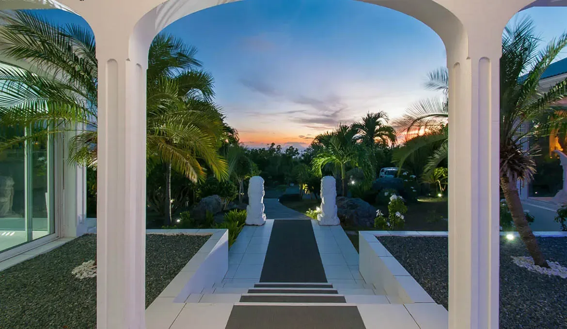 Experience the magic of St. Barts at Villa Byzance, where the elegant archway frames a breathtaking sunset over the lush tropical landscape. Your journey begins along a serene pathway lined with palm trees and illuminated sculptures, leading to the luxury and tranquility of this exclusive villa retreat.                                                                                                                                                                                                         +
 