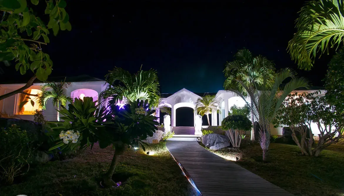 Step into a tropical sanctuary at Villa Byzance, where a glowing wooden pathway winds through vibrant gardens toward the villa's elegant, softly lit entrance under a star-dusted Caribbean night. Immerse yourself in the serenity of St. Barths as illuminated palms and floral accents frame your arrival at this luxurious retreat.                                                                                                                                                                               +
 