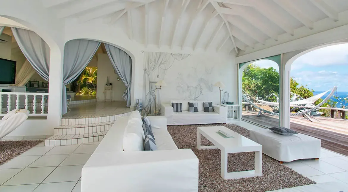 Step into the serene elegance of Villa Byzance's living room, where crisp white linens and airy arches frame uninterrupted ocean vistas. Let the gentle breeze carry you from plush sofas to the hammock-dotted deck, where St. Barths' turquoise waters and lush hillsides create a private paradise.                                                                                                                                                                                                      +
 