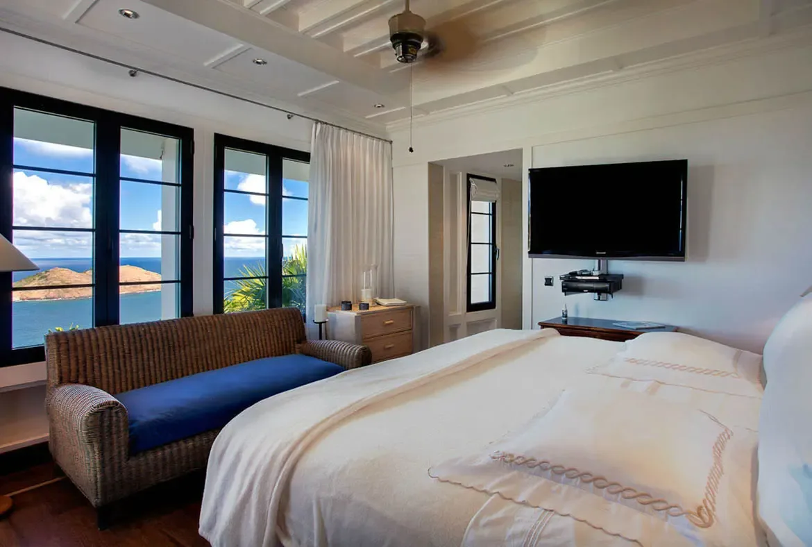 Wake to the Caribbean horizon framed by floor-to-ceiling windows. A stylish blue settee by the glass creates a private nook for morning coffee, while crisp white linens and vaulted ceilings evoke unhurried elegance in your hillside sanctuary.