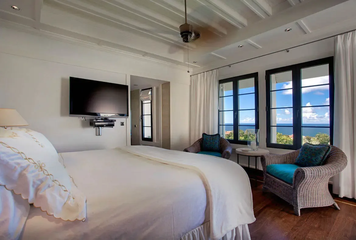 Dark-framed windows frame the Caribbean horizon like living paintings. A private sitting nook with wicker chairs invites morning coffees by the ocean, while coffered ceilings and crisp white linens create a serene, airy sanctuary.