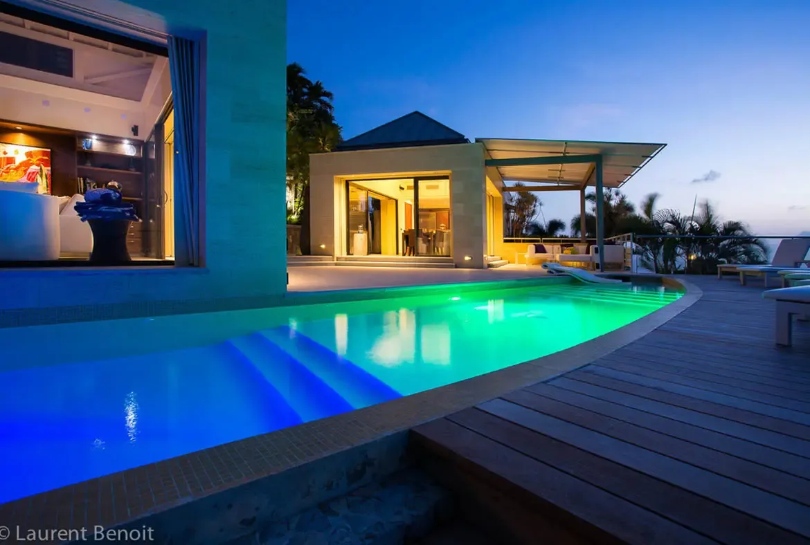 During the magical blue hour, floor-to-ceiling glass doors vanish to merge living room and pool deck. The LED-illuminated infinity pool transitions from cobalt to turquoise, while warm interior lighting creates a welcoming lantern effect against the twilight.