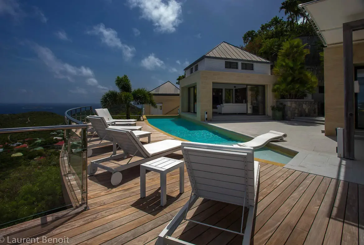 Bask in brilliant natural light on the expansive hardwood deck, where a crystalline lap pool reflects the Caribbean sky. Limestone masonry and classic gabled roofs frame panoramic views across emerald hillsides to the turquoise horizon.