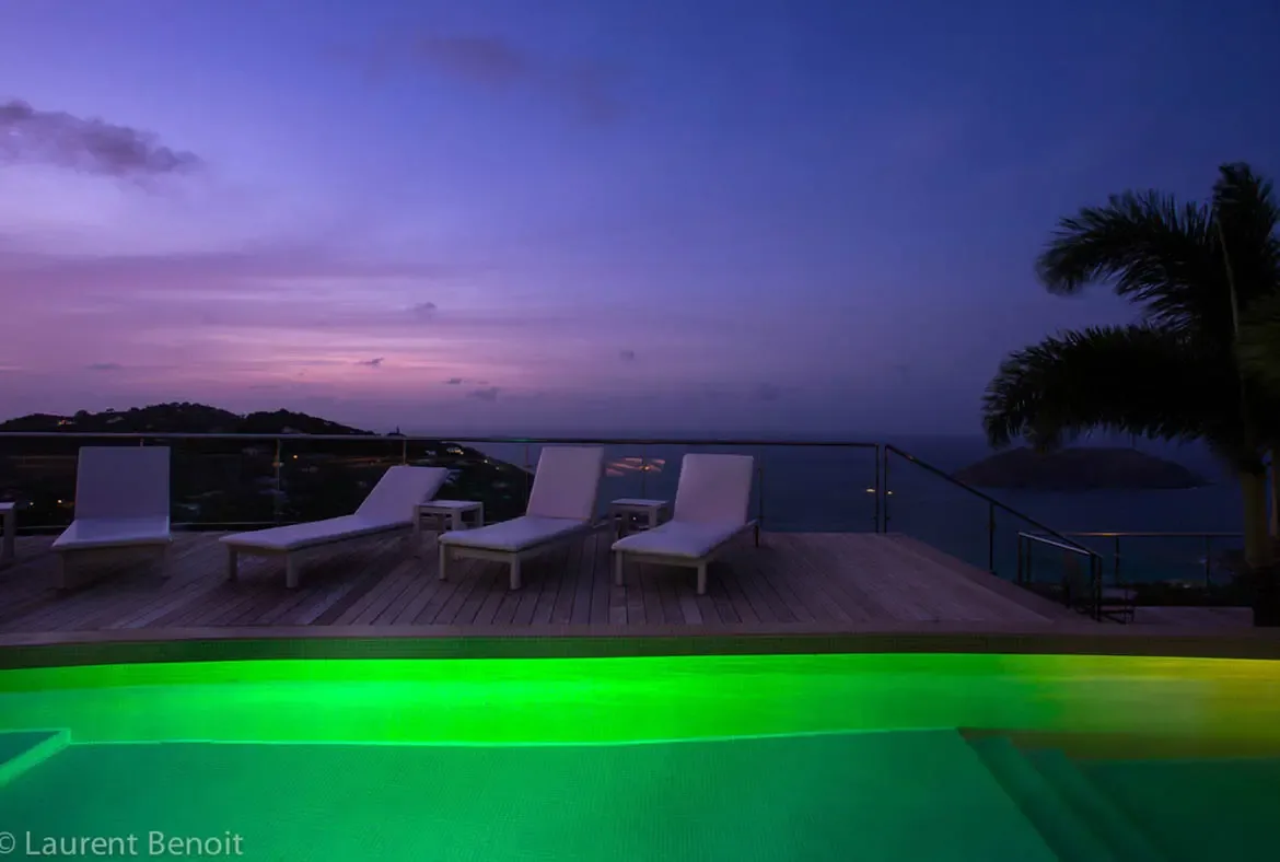 The neon-emerald glow of the infinity pool contrasts against a velvet purple twilight. Perched high above the coastline, the expansive deck offers 180-degree views of darkening ocean and twinkling town lights—a front-row seat to Caribbean magic.