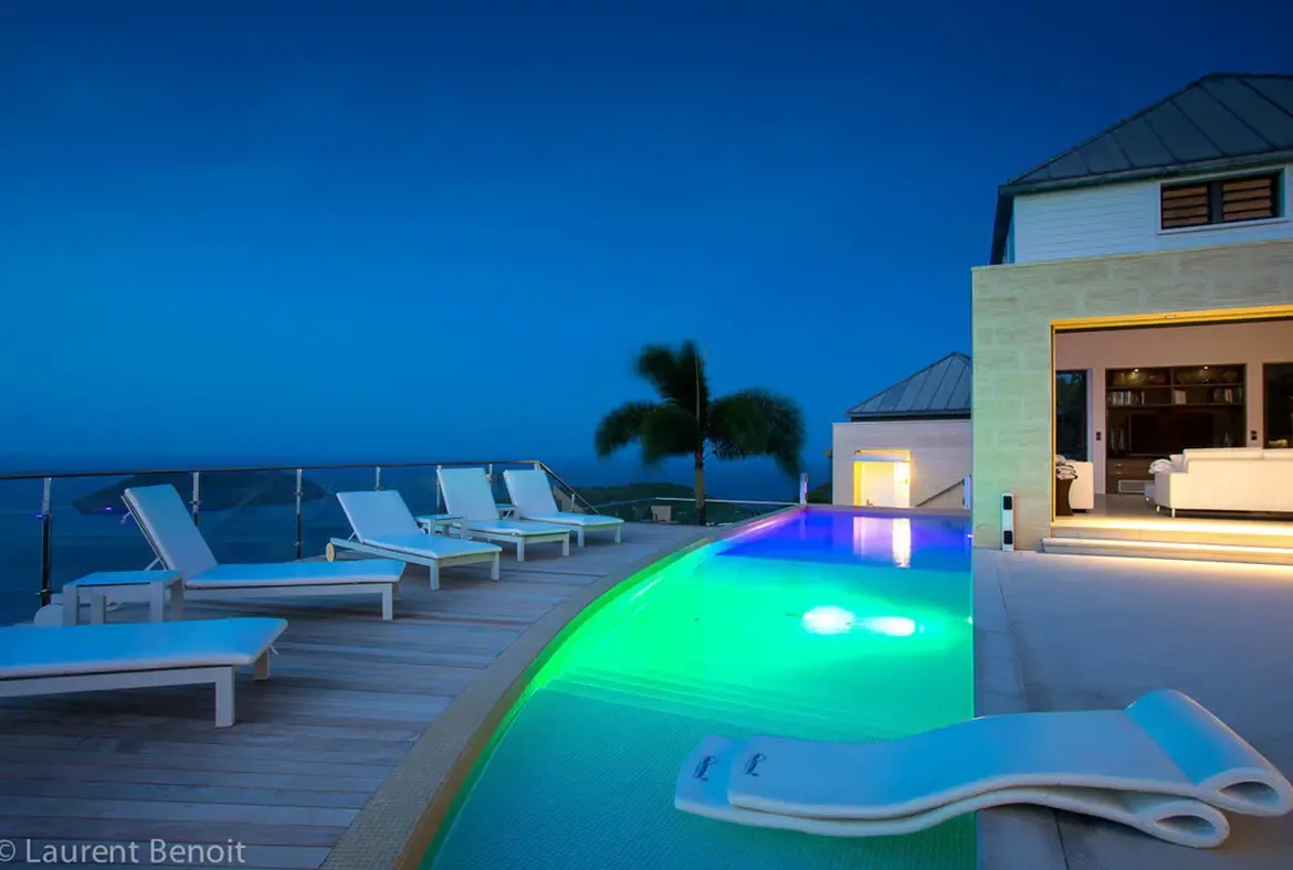 The curved infinity pool glows like a sapphire gem against the indigo sky. A glass-railed sundeck offers 180-degree ocean views, while the open-plan living area flows seamlessly onto the deck—pure escapism perched high above the Caribbean.