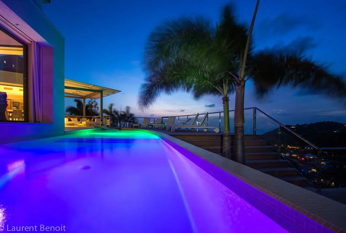 The infinity pool transitions from deep purple to neon green as twilight deepens. Glass railings frame panoramic views of twinkling valley lights below, while a covered patio offers warm interior comfort.