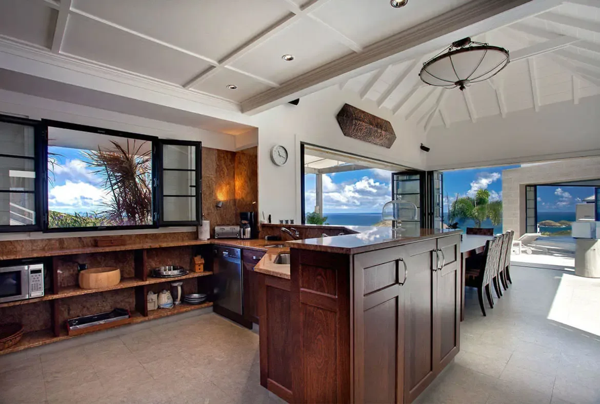 The gourmet kitchen becomes a social hub with folding glass doors that erase boundaries between chef and scenery. Vaulted ceilings with exposed beams crown the dual-level island, where meal prep becomes a shared event against turquoise waters.