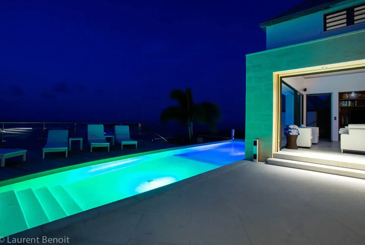 A long lap pool glows with neon-green light against the deep indigo sky. Floor-to-ceiling glass doors vanish to merge the bright living room with the stone patio, while teal loungers line the deck.