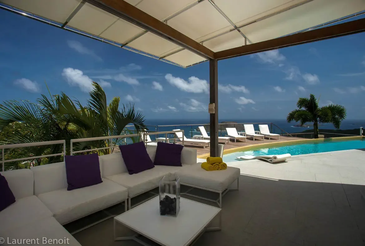 Relax on the plush white sectional with deep amethyst accents, where a contemporary canopy shades your outdoor living area. The infinity pool merges with the Caribbean horizon, and a submerged lounge chair offers the ultimate way to stay cool while soaking in the view.