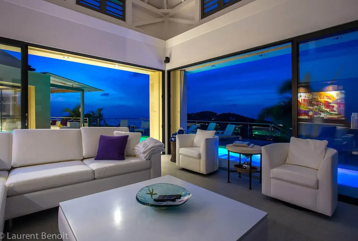 Floor-to-ceiling glass vanishes to merge your living space with the Caribbean twilight. The cobalt sky matches the illuminated pool, while all-white furnishings create a cloud-like atmosphere against the vaulted pavilion ceiling.