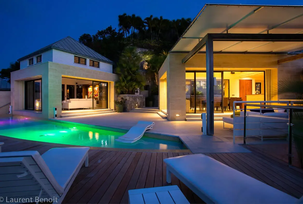 A private luminous sanctuary where golden interior glow contrasts with emerald pool lighting. Separate pavilions connected by stone pathways offer various zones for relaxation—from sun-drenched decks to shaded pergolas—in your own private resort.
