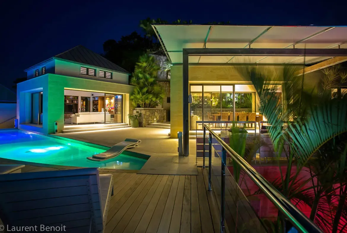 Vibrant emerald LED lighting transforms the pool into a theatrical centerpiece. Two-story and single-level pavilions glow with warm interior light, while the wooden deck invites evening lounging under tropical palms.