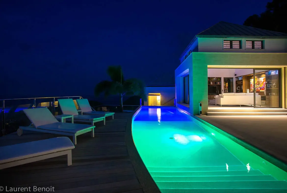 The villa transforms into a glowing jewel box at night. Emerald and sapphire underwater lighting creates an immersive focal point, while warm golden interiors visible through floor-to-ceiling glass sliders invite you to blur the line between indoor comfort and fresh evening air.