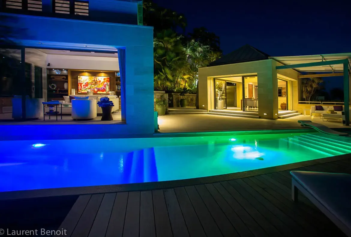 The pool glows with vibrant LED lighting—deep blue and emerald green—seen from the wooden deck. Two illuminated pavilions frame the scene: the main living area with bold artwork to the left, and a guest pavilion to the right.