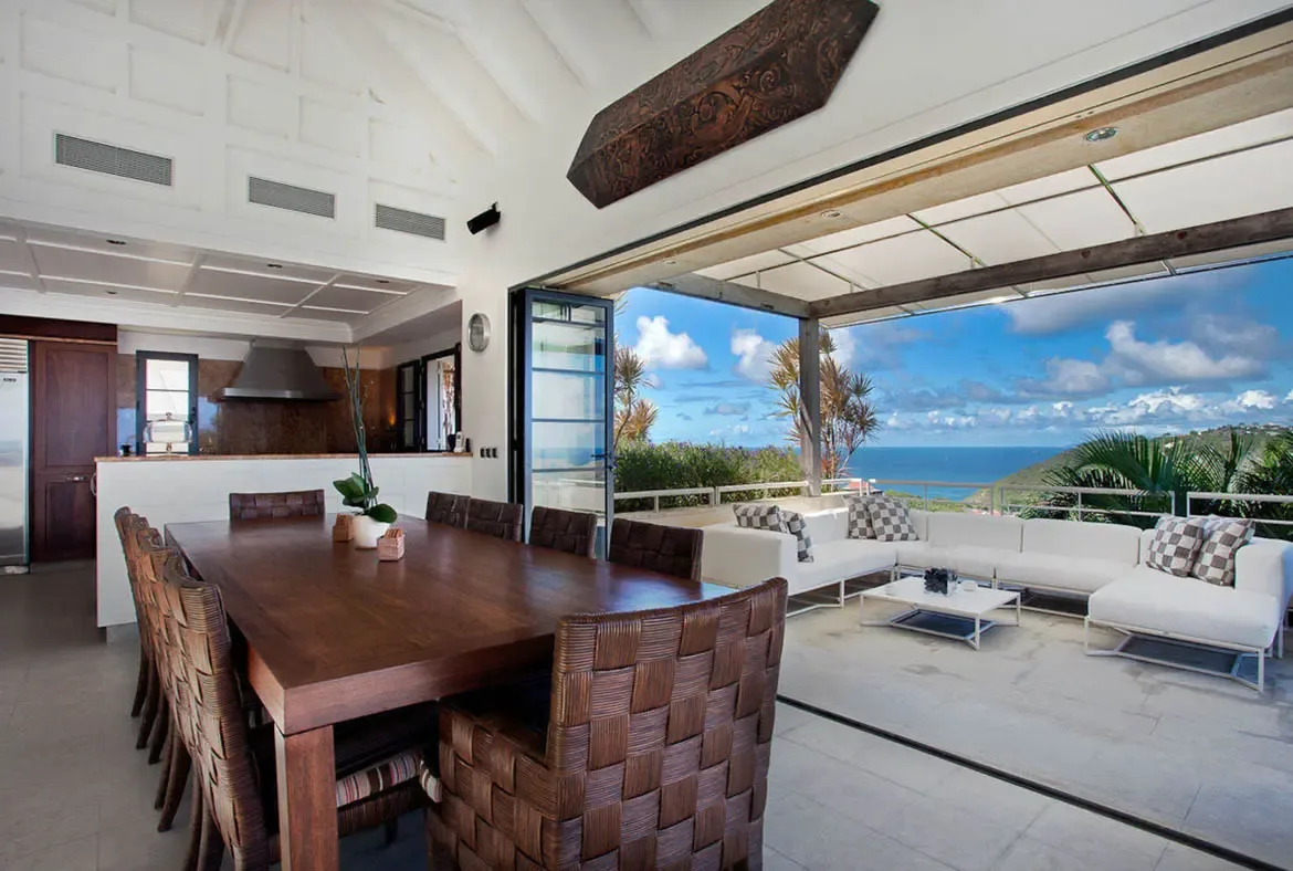 Massive folding glass doors disappear to merge dining and living spaces with the Caribbean horizon. A rich wood dining table and hand-woven chairs anchor the vaulted white ceilings, creating the ultimate entertaining hub with cinematic ocean views.