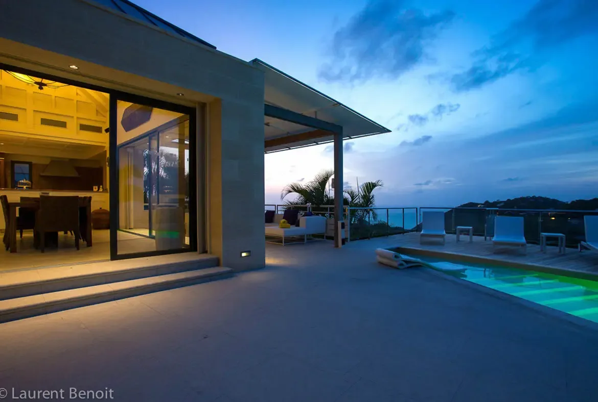 As twilight settles, warm interior lighting creates a golden contrast against the luminous green pool. Oversized sliding doors transform the living area into an open-air pavilion, blending gourmet comfort with Caribbean serenity.