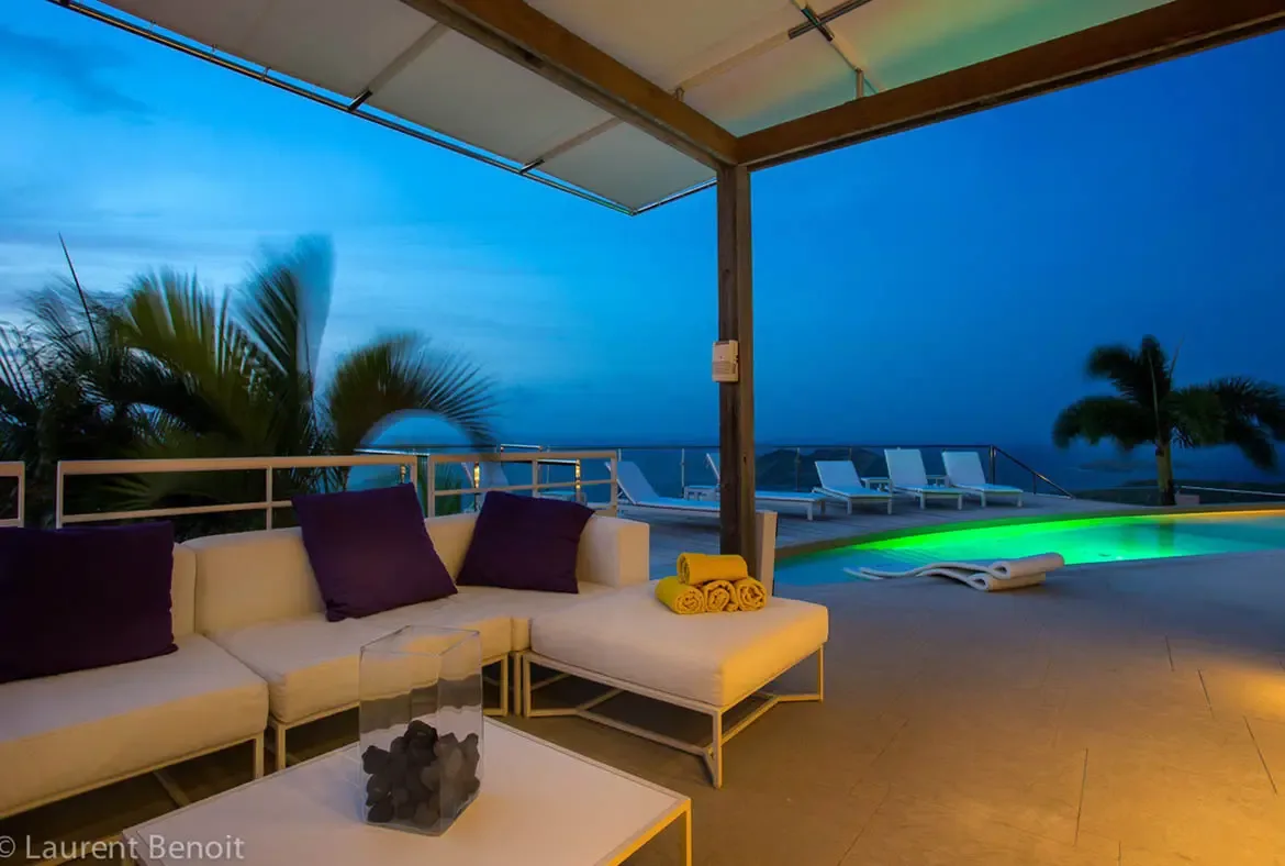 The neon-green glow of the infinity pool creates a striking centerpiece for evening entertaining. A cantilevered roof shades the designer white sectional with purple accents, while glass railings ensure your ocean panorama remains uninterrupted.