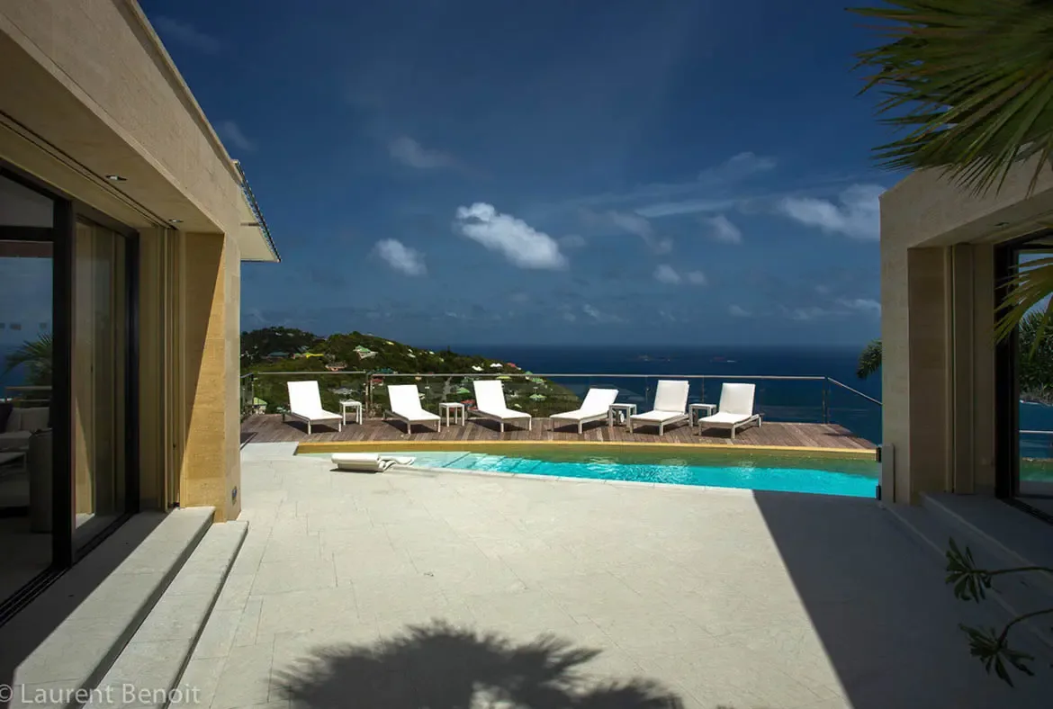 The infinity pool spills into the deep blue Caribbean from this hilltop sanctuary. Floor-to-ceiling glass walls disappear into limestone architecture, while six white chaises invite sunbathing with panoramic views of rolling hillsides and distant islands.