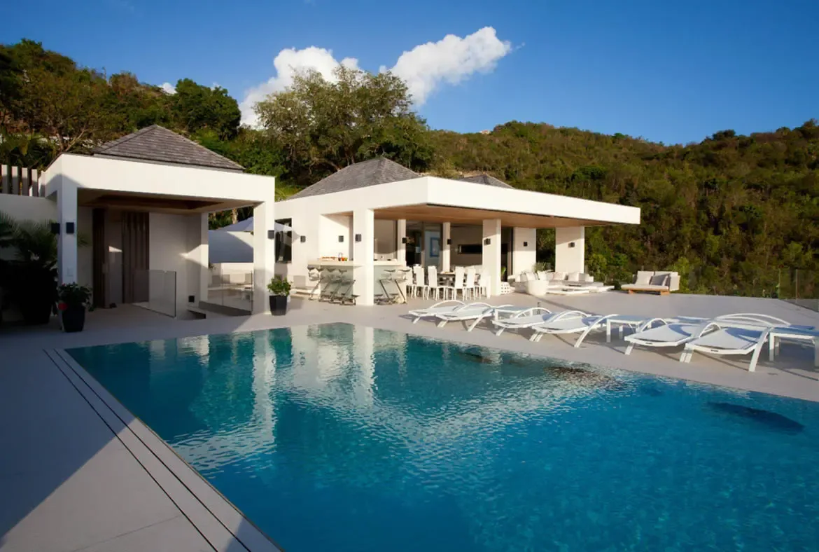 A modern white villa with two pavilions perches on a lush hillside, where a turquoise infinity pool, white loungers, and an outdoor bar and dining area overlook a green valley beneath a bright blue sky.