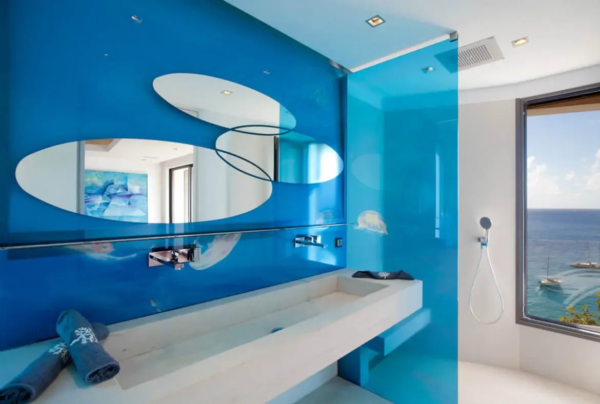 A glossy blue accent wall with three overlapping oval mirrors and jellyfish motifs sits above a stone trough sink, where a glass-partitioned rain shower opens to an ocean view with a white sailboat.