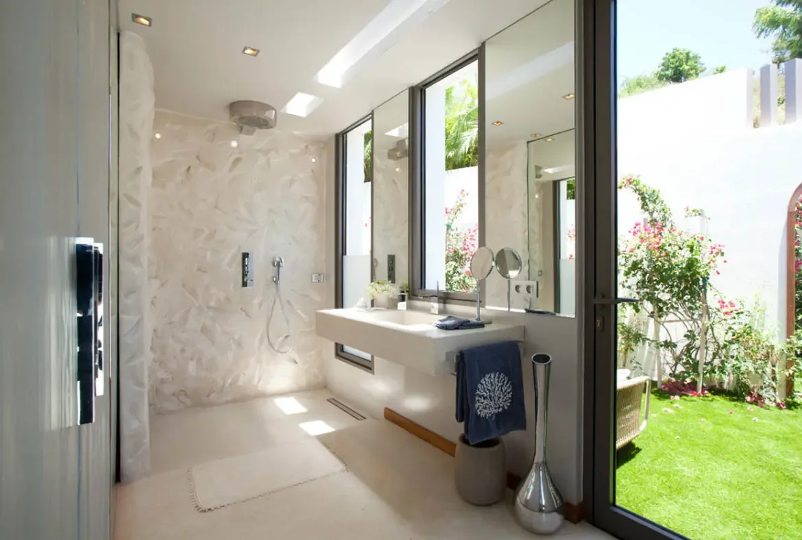 A white floating vanity with an integrated sink sits beneath a large mirror, where an open-concept rain shower with shell-patterned tiles opens to a private garden with pink bougainvillea.