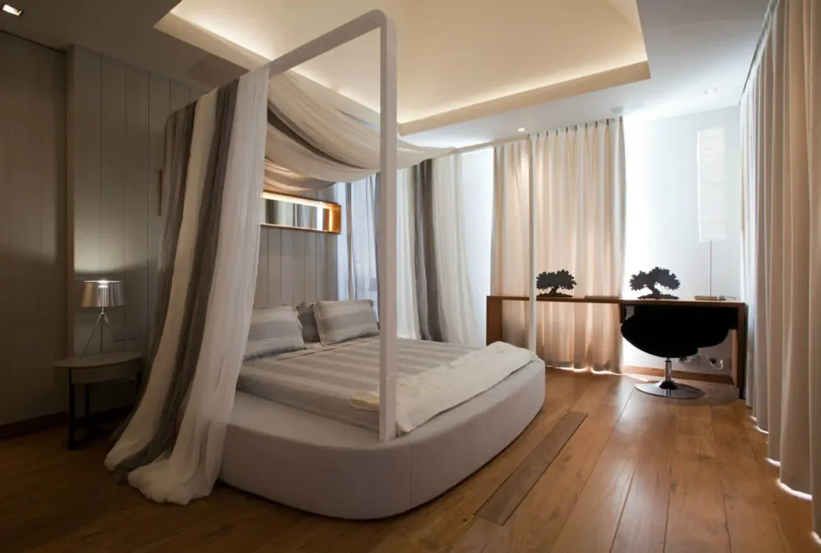 An oval platform bed with sheer white-and-grey canopy drapes sits beneath cove-lit ceilings, where a slim wooden desk with black tree silhouettes and a floor lamp accent the serene neutral space.