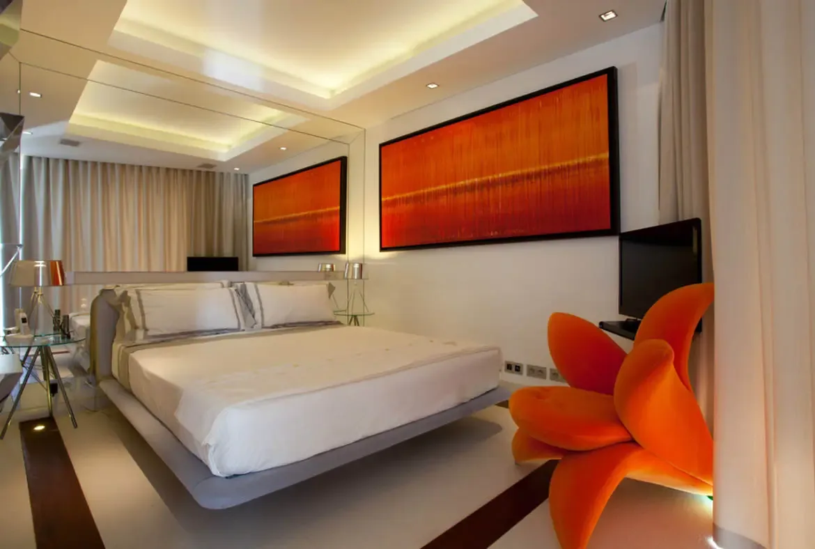 A low platform bed before a floor-to-ceiling mirrored wall sits beneath cove-lit ceilings, where a vibrant orange abstract painting and a sculptural orange flower chair add bold color to the neutral space.