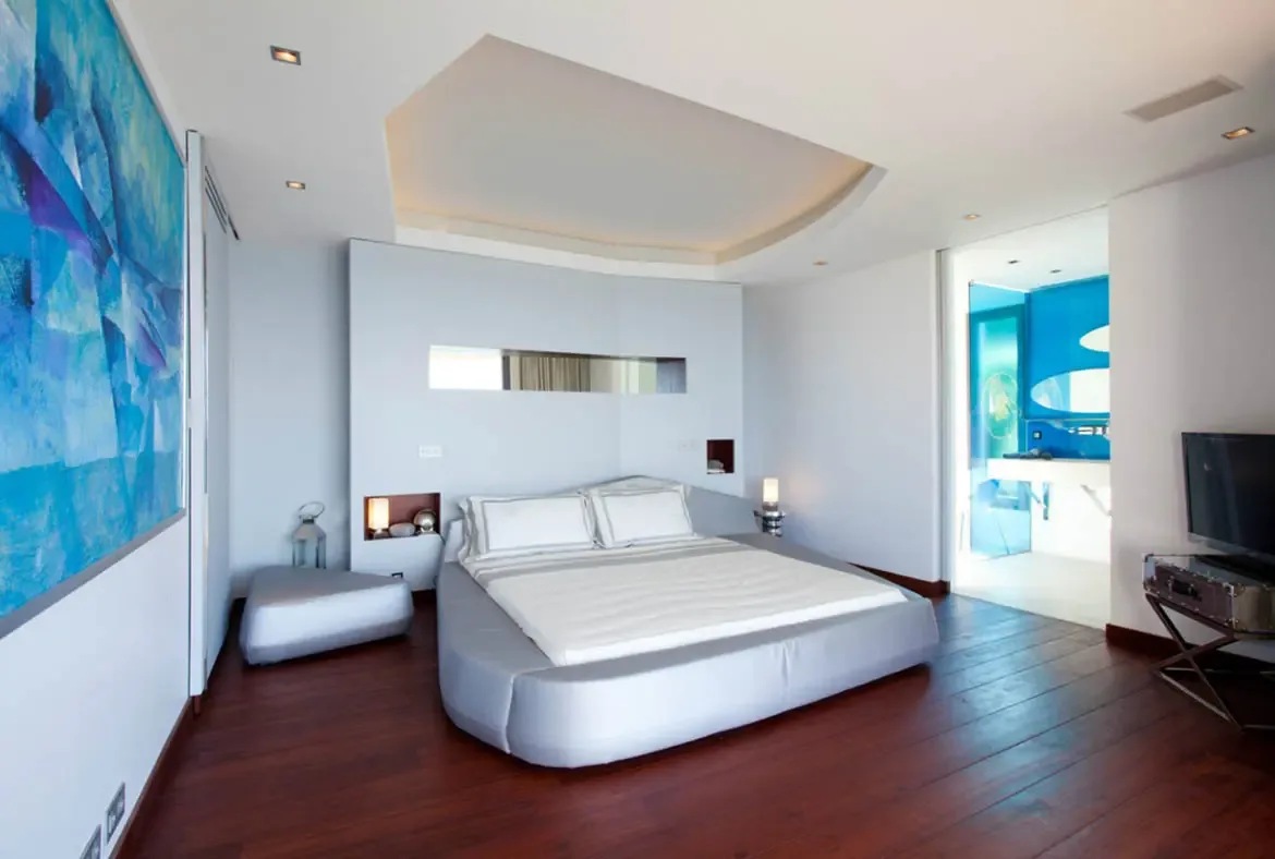 An oval platform bed with grey linens sits beneath a vibrant blue abstract painting, where a mirrored niche and cove-lit ceiling accent a dark wood floor and a blue-accented bathroom glimpses beyond.