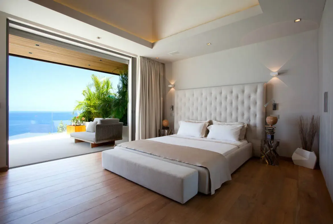 A tall tufted grey headboard with white linens and a bench faces open glass doors, where a stone terrace daybed and a panoramic ocean view invite seamless indoor-outdoor living.