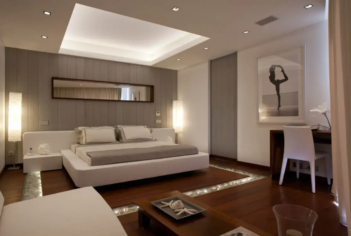 A white platform bed over illuminated river stones in a glass trench sits beneath a mirrored wall and cove-lit ceiling, where starfish on a wooden tray and a black-and-white dance photo accent the room.