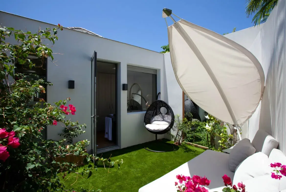 A white outdoor daybed and a black hanging egg chair sit on artificial green grass, where bright pink bougainvillea spills over white walls beneath a leaf-shaped sunshade and a clear blue sky.