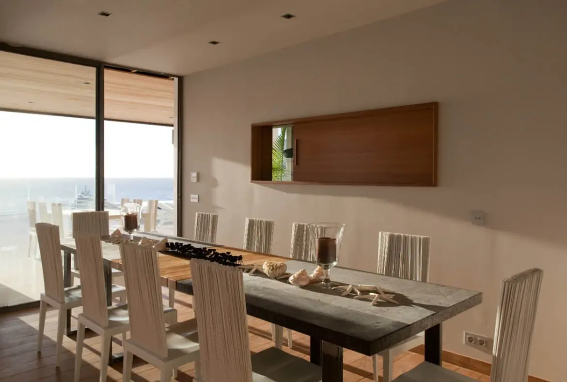 A dark stone dining table with white cord-strung chairs and seashell decor faces floor-to-ceiling glass walls, where a bright coastal panorama fills the room with natural light.