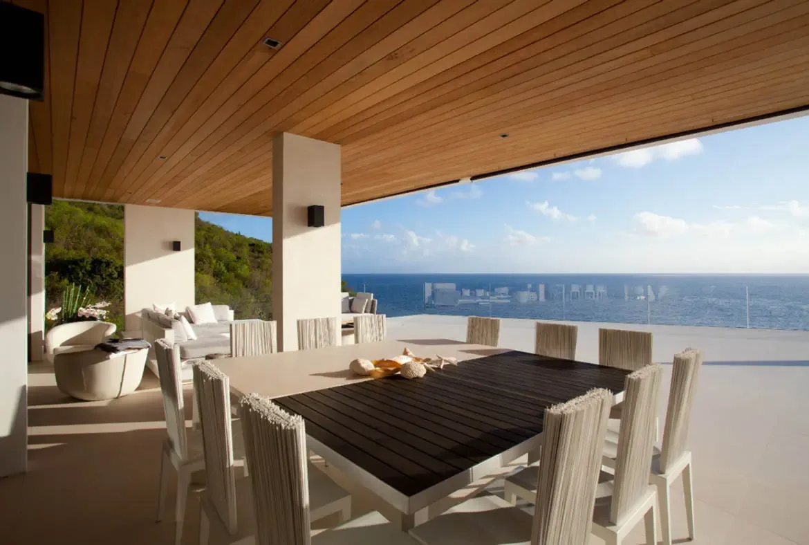 A long dining table with white cord chairs and seashell decor sits beneath a warm wood ceiling, where white columns and a glass railing frame a panoramic deep blue ocean under a bright sky.