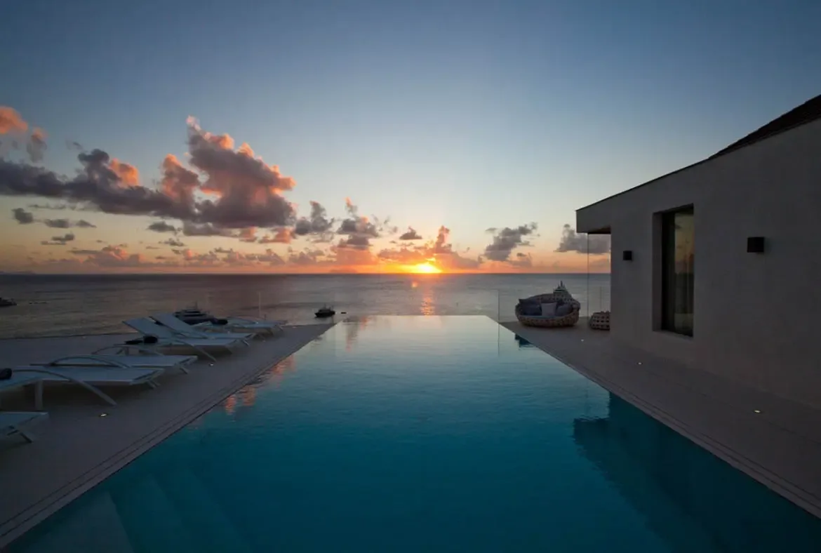 An infinity pool mirrors a golden sunset between white loungers and a woven bowl chair, where the sun casts a shimmering path across calm ocean water beneath wispy orange-tinted clouds.