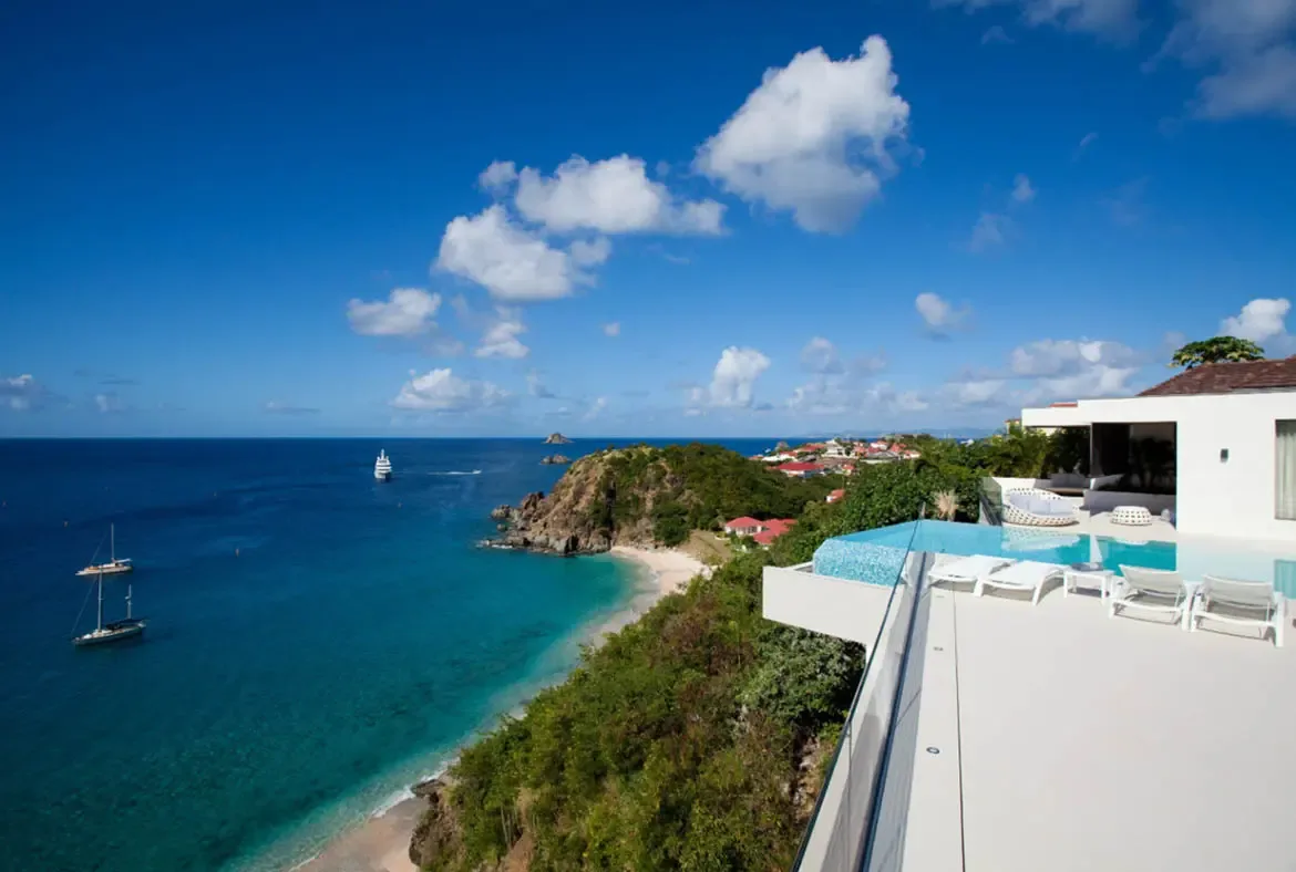 An infinity pool on a white terrace overlooks a turquoise bay with a cruising yacht, where white loungers and a circular wicker daybed frame a secluded beach cove beneath lush green hills.