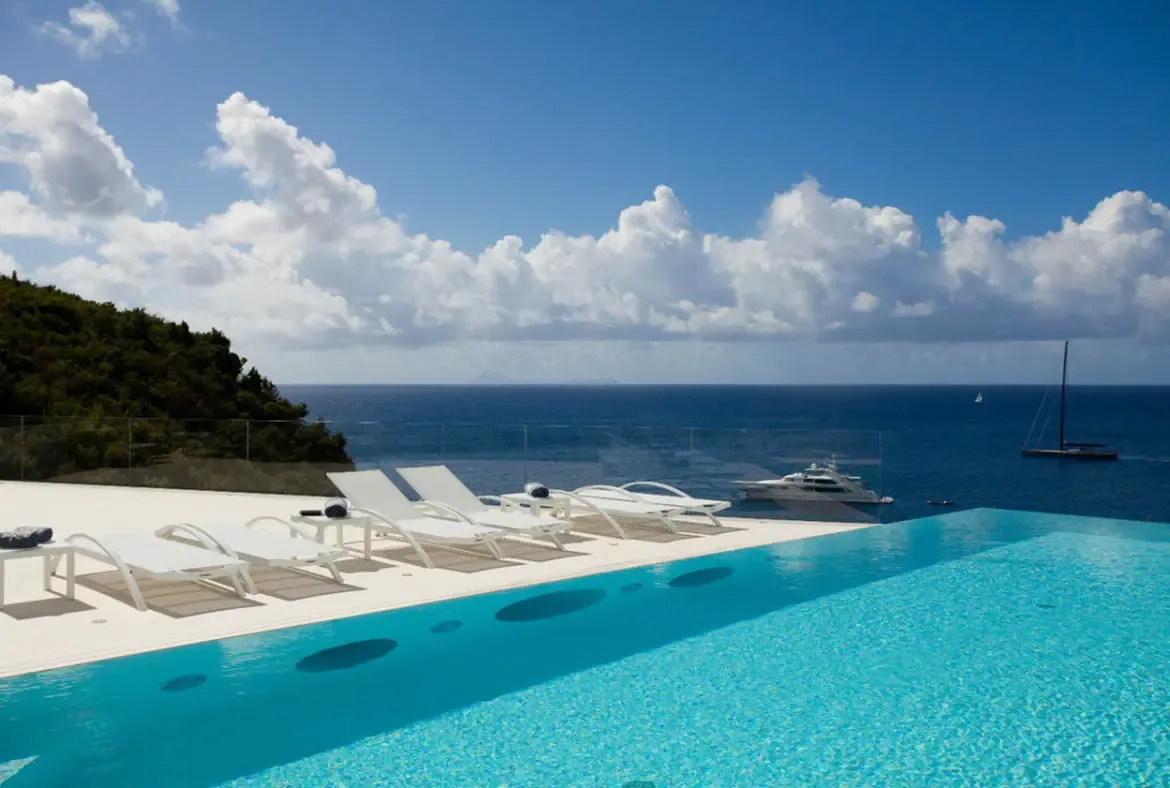 An infinity pool with turquoise water sits beside white loungers on a minimalist deck, where a glass railing opens to a deep blue bay with anchored yachts and sailboats beneath puffy cumulus clouds.