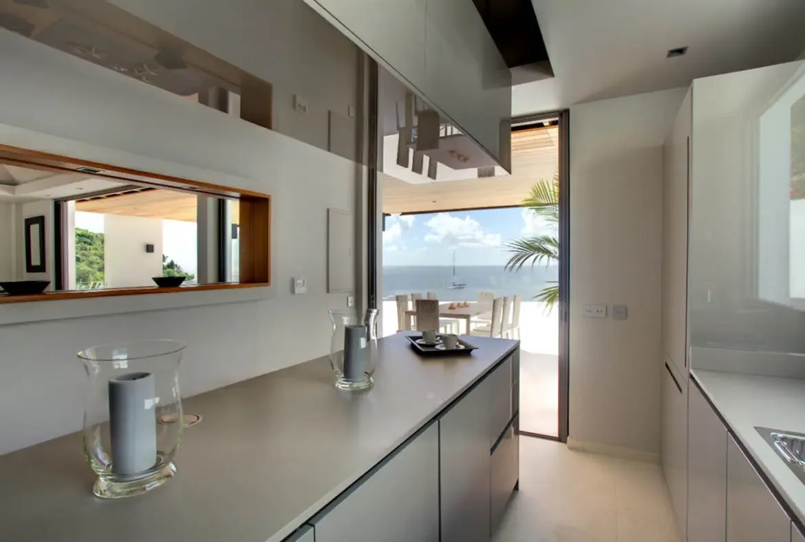 A matte grey kitchen island with glass candle lanterns sits beneath reflective cabinetry, where a wide glass doorway opens to a sun-drenched terrace and a calm blue ocean under wispy clouds.