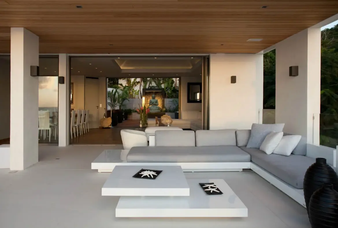 An L-shaped grey sofa with blue pillows sits before open glass walls, where a stone Buddha statue framed by red heliconia flowers and tropical greenery creates a tranquil Zen courtyard focal point.