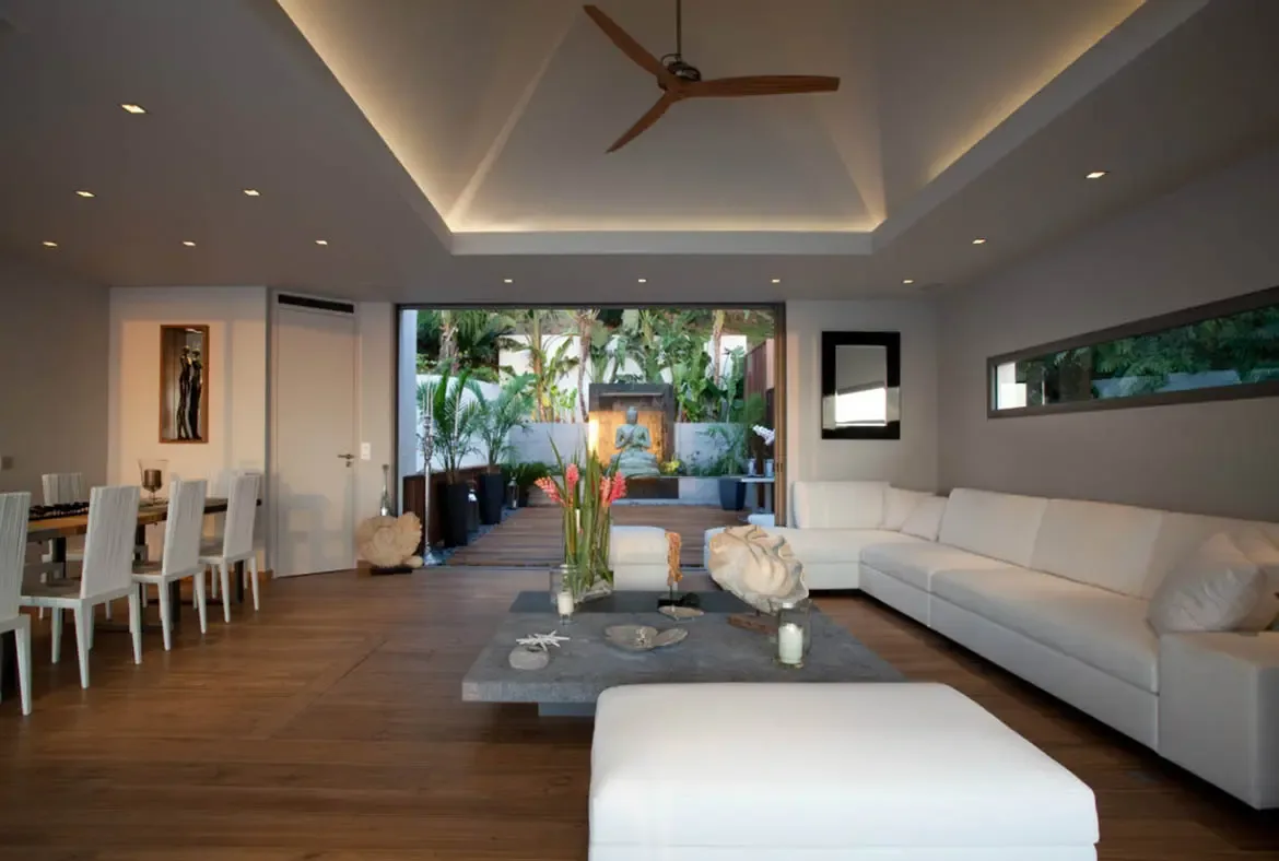 A white leather L-shaped sofa with a grey stone coffee table and tropical flowers sits before open glass walls, where a stone Buddha in a lush garden courtyard brings serenity to the open-concept space.