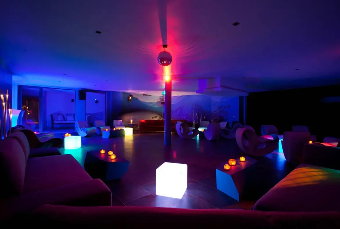 A moody lounge with purple sofas and LED cube stools glows beneath blue and pink ambient light, where a disco ball and a marine-themed mural with fish silhouettes create a private nightclub atmosphere.