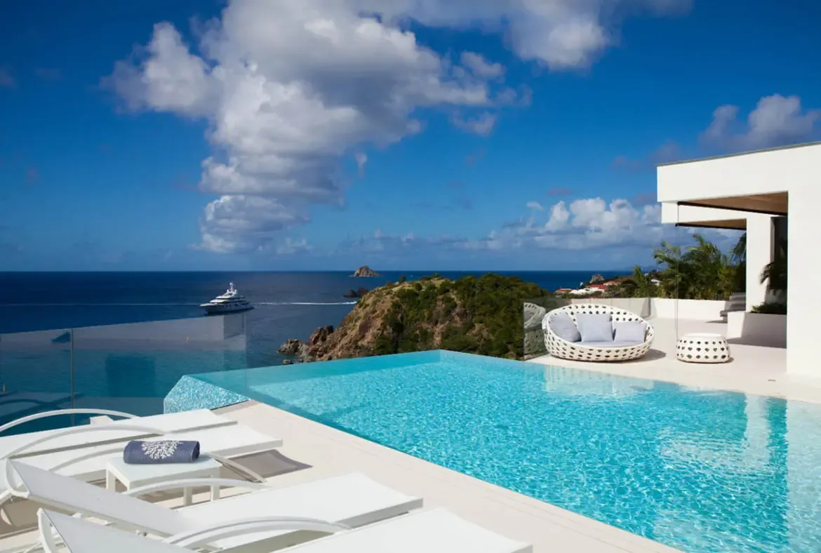 White loungers beside an infinity pool and a circular wicker daybed face a deep blue sea, where a white yacht cruises beneath puffy cumulus clouds above a lush green headland.