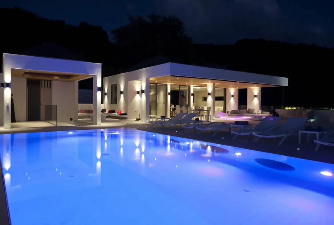A glowing blue pool reflects a white villa with dramatic hourglass wall sconces at night, where grey loungers and outdoor sofas with red pillows sit beneath warm-lit eaves against a dark tropical hillside.