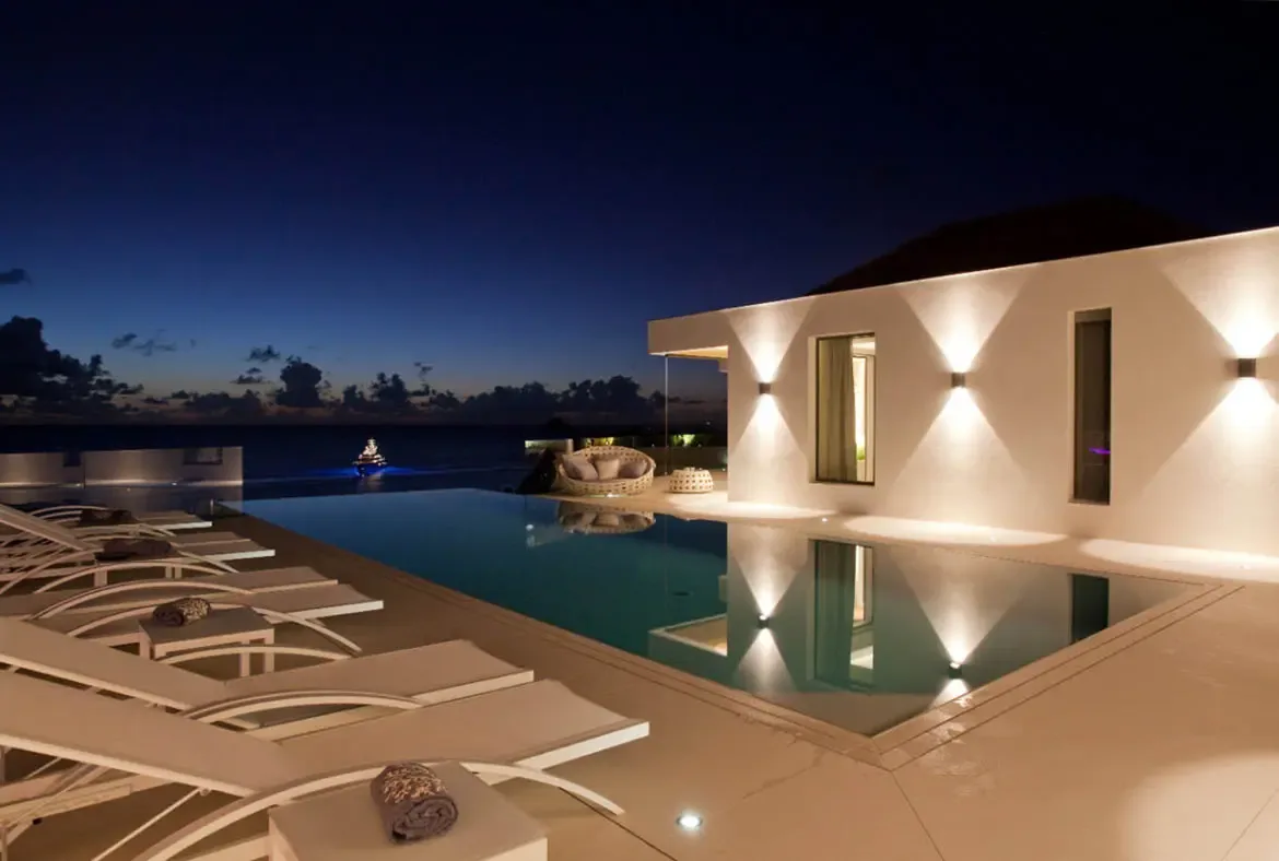 An infinity pool reflects a white villa with hourglass wall sconces at twilight, where white loungers and a woven daybed line the deck beneath an indigo sky with a lingering orange glow on the horizon.