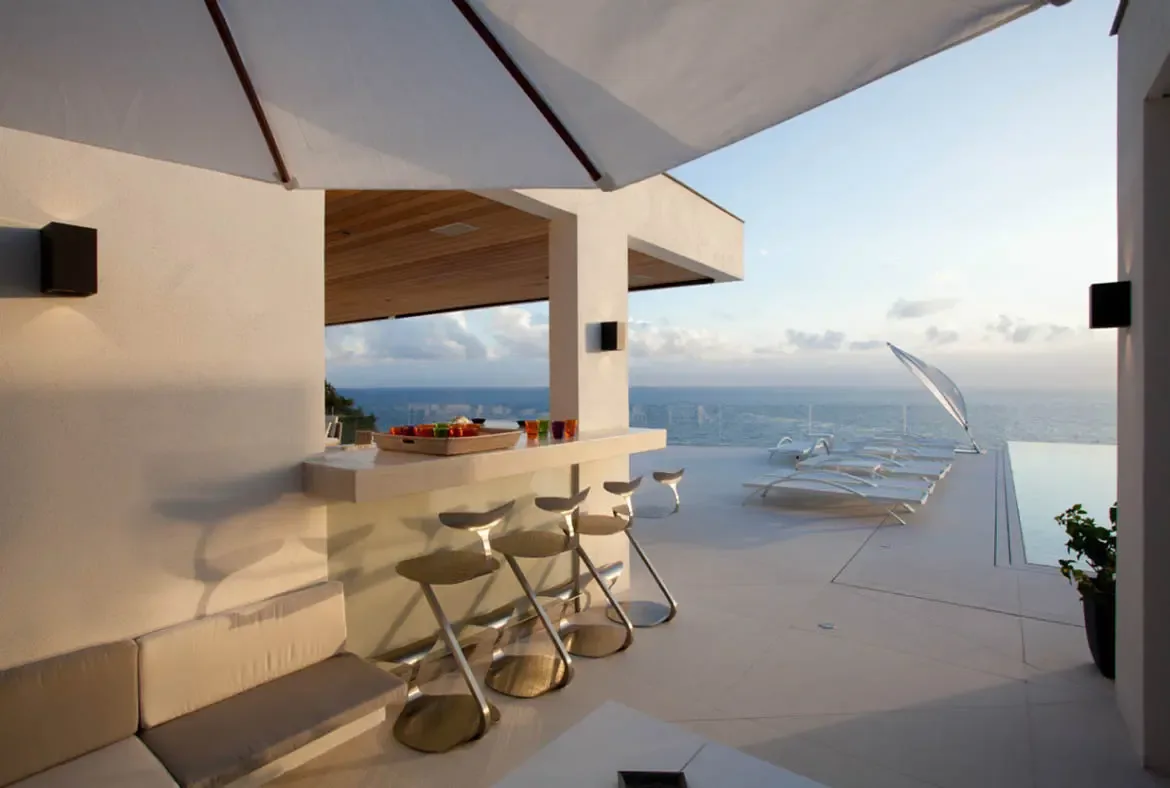 A white stone bar with sculptural stools and colored glassware sits on a terrace, where grey modular sofas and white loungers face an infinity pool and a calm ocean beneath a warm golden-hour sky.