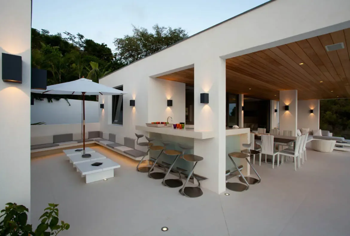 A white cantilevered bar with sculptural stools and a long dining table under a wood-plank ceiling glow at dusk, where wall sconces cast warm hourglass light and lush greenery rises behind white walls.