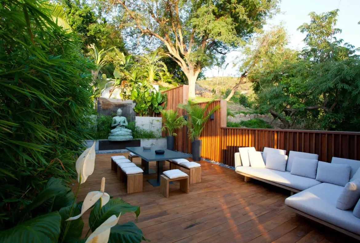 A charcoal dining table with white-cushioned stools sits on a wood deck beside a stone Buddha in a garden alcove, where bamboo and banana trees create a Zen-inspired private retreat.