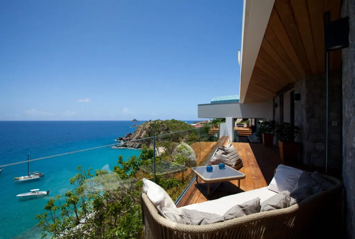 A curved outdoor sofa with grey pillows sits on a wood deck beside a glass railing, where a turquoise bay with anchored sailboats and lush green hills stretches beneath a clear blue sky.