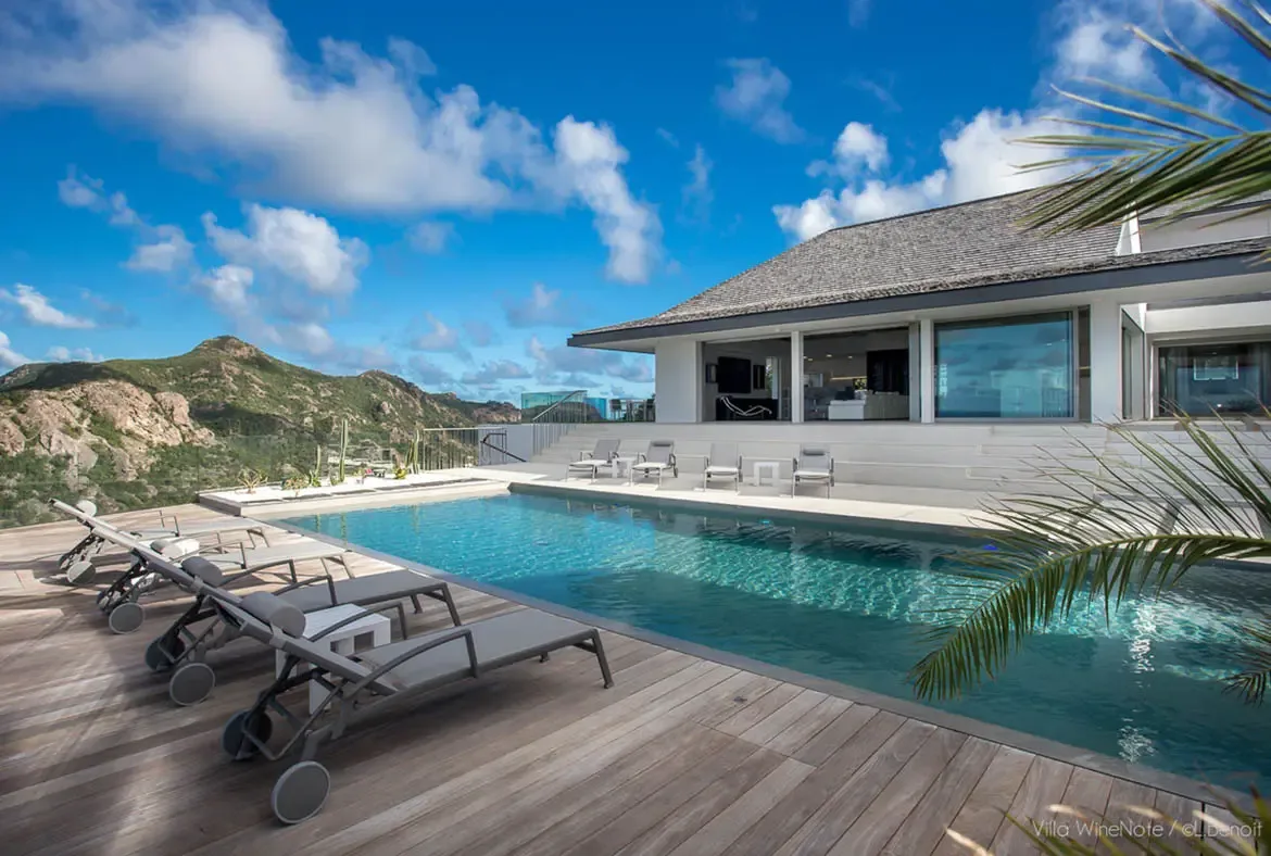 An infinity pool with grey loungers sits before a white modern villa with open glass doors, where cactus gardens and rolling green mountains stretch beneath a bright blue sky.
