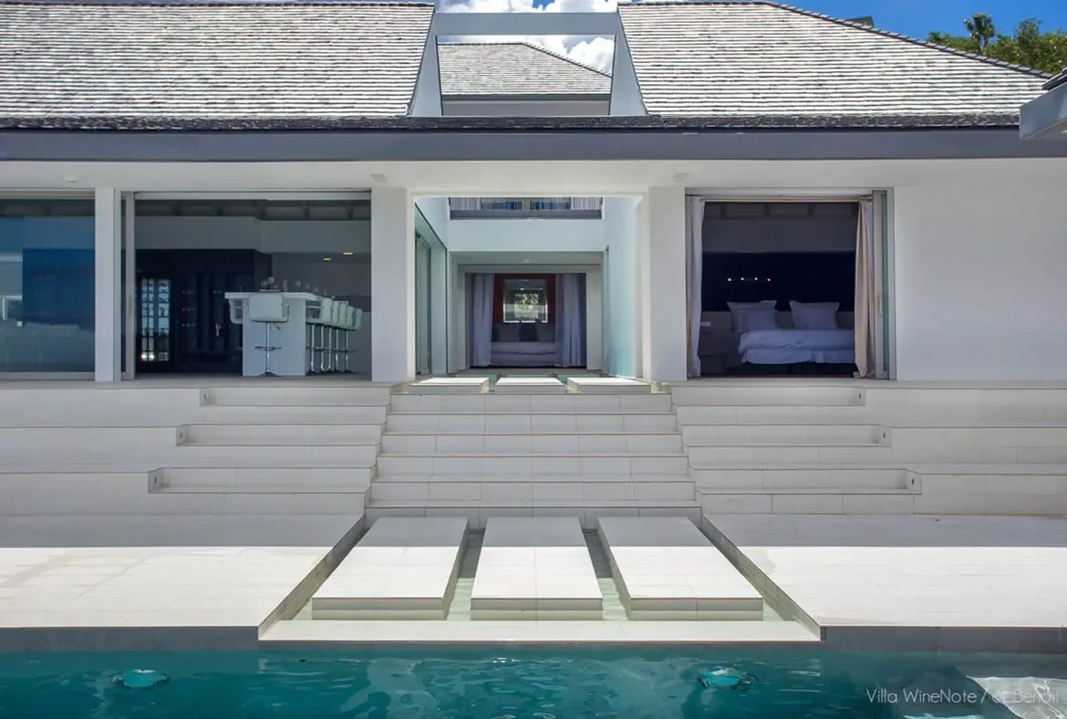 A turquoise reflection pool with white stone stepping stones leads to tiered white steps, where a white villa with open living spaces and a wine display frames a panoramic ocean view.